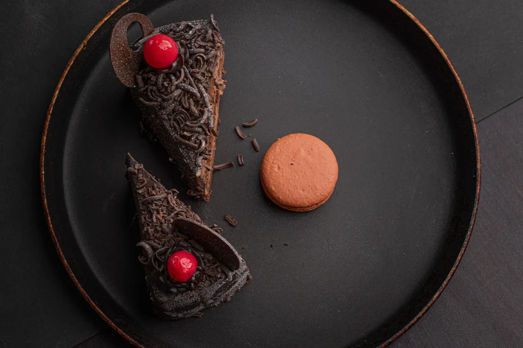 Two slices of chocolate cake with red cherries and a macaron on a black platter.