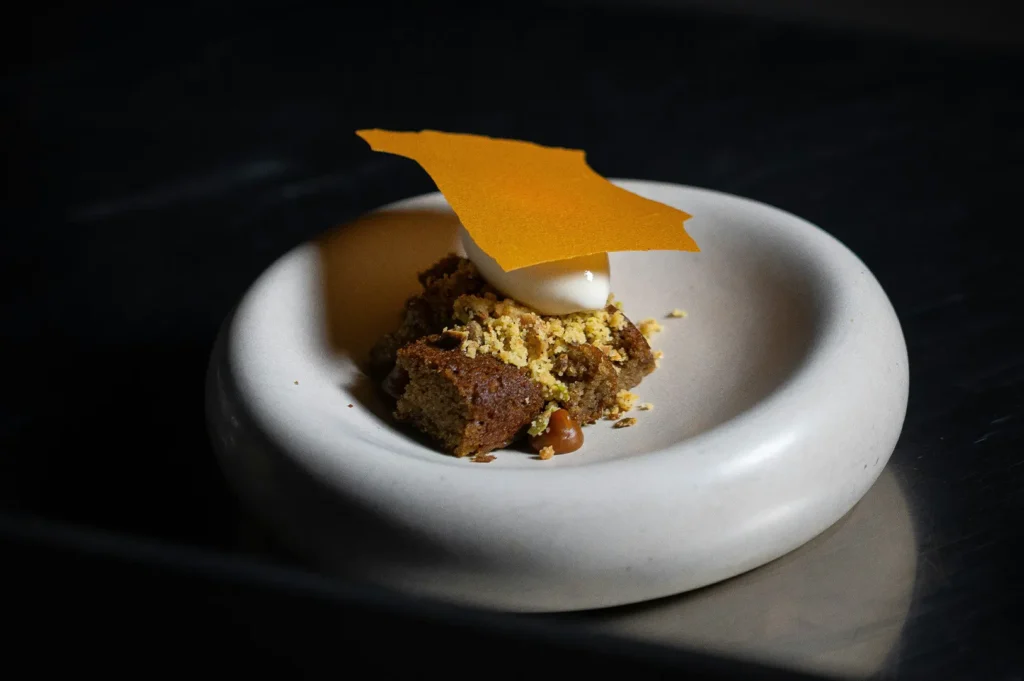 Plated gourmet dessert featuring cake, crumble, ice cream, and an orange crisp in a modern white bowl.