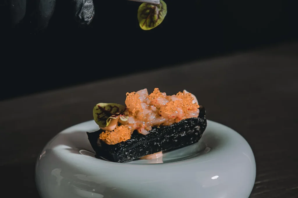 Black sushi roll topped with orange tobiko and green microgreens on a white plate.
