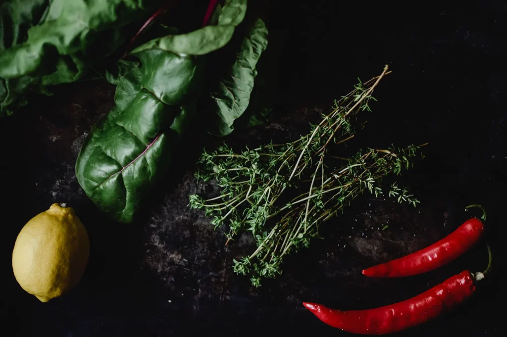 Dark moody food photography featuring fresh leafy greens, sprigs of thyme, a bright yellow lemon, and red chili peppers on a black textured background.