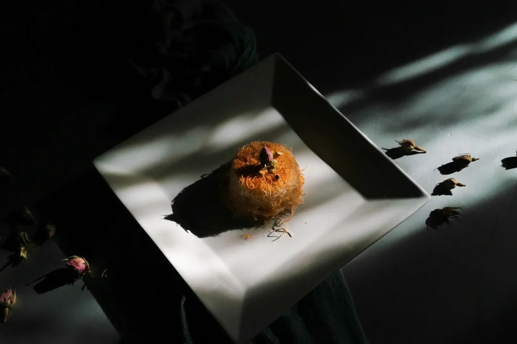 A single piece of golden-brown kunafa topped with a dried rosebud, served on a square white plate with moody, dramatic lighting.