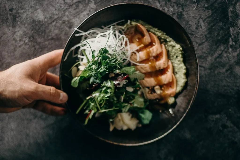 A hand holding a dark ceramic bowl featuring a fresh seared salmon salad with greens, radish, and dressing on a dark rustic background.