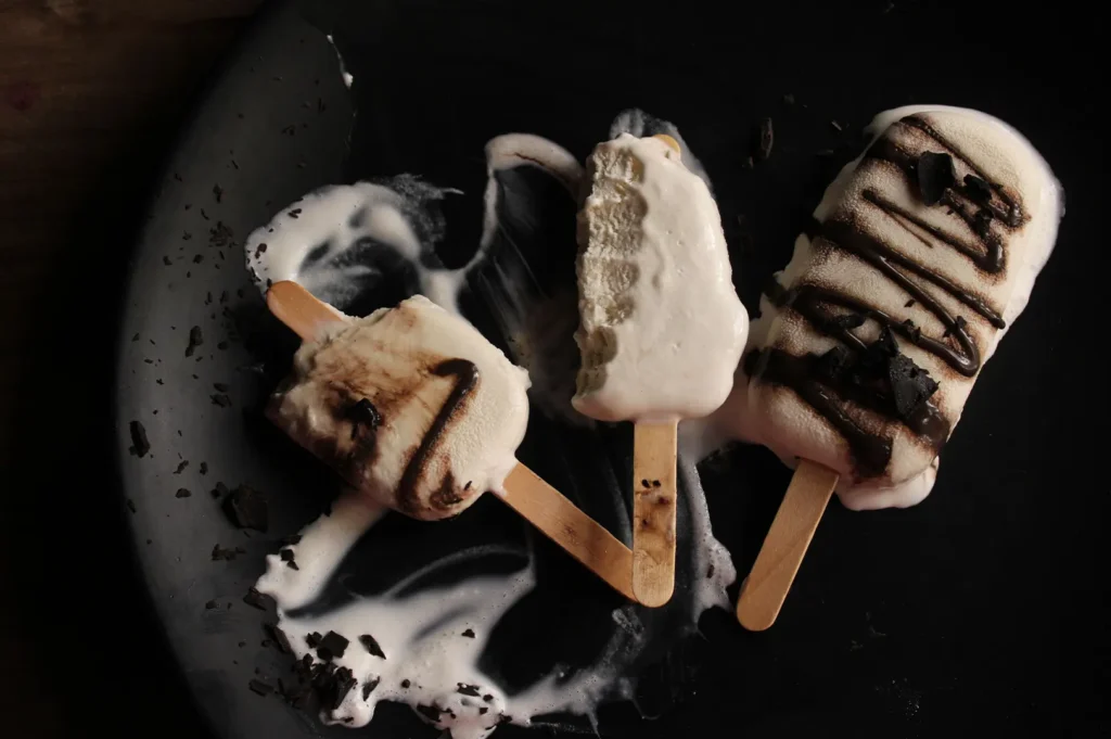 Three creamy vanilla ice cream bars drizzled with chocolate syrup and chocolate shavings on a dark plate.