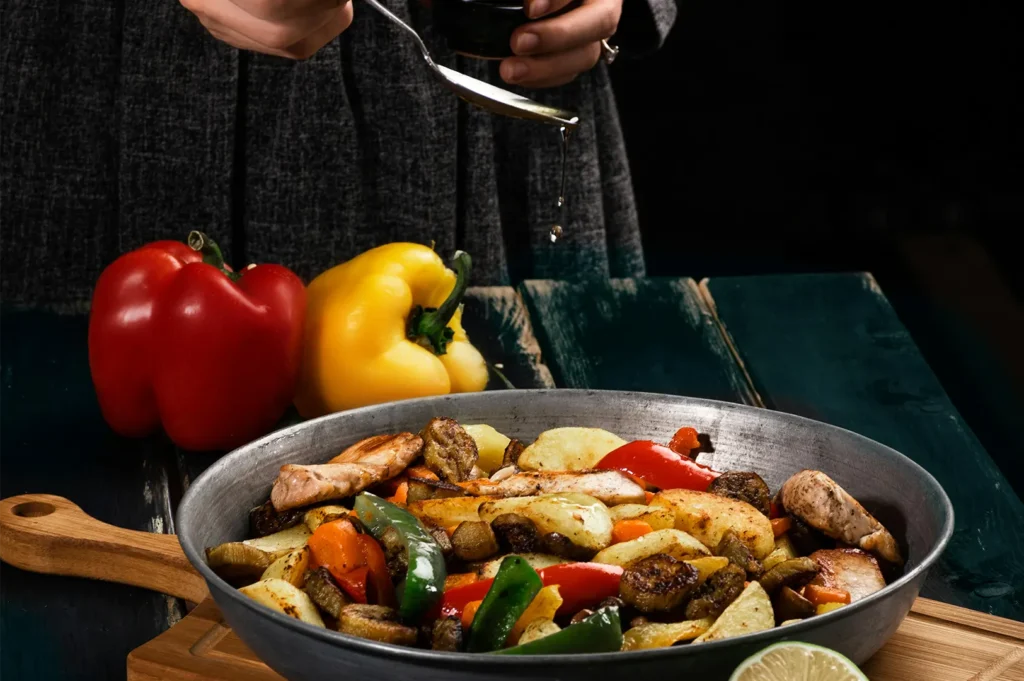 Skillet of cooked vegetables and chicken with a cook drizzling oil, next to fresh bell peppers.