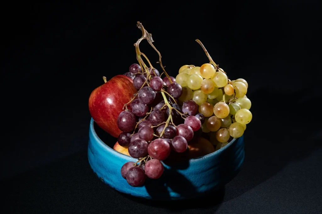 Fresh red apple, red grapes, and green grapes in a blue ceramic bowl on a dark background.