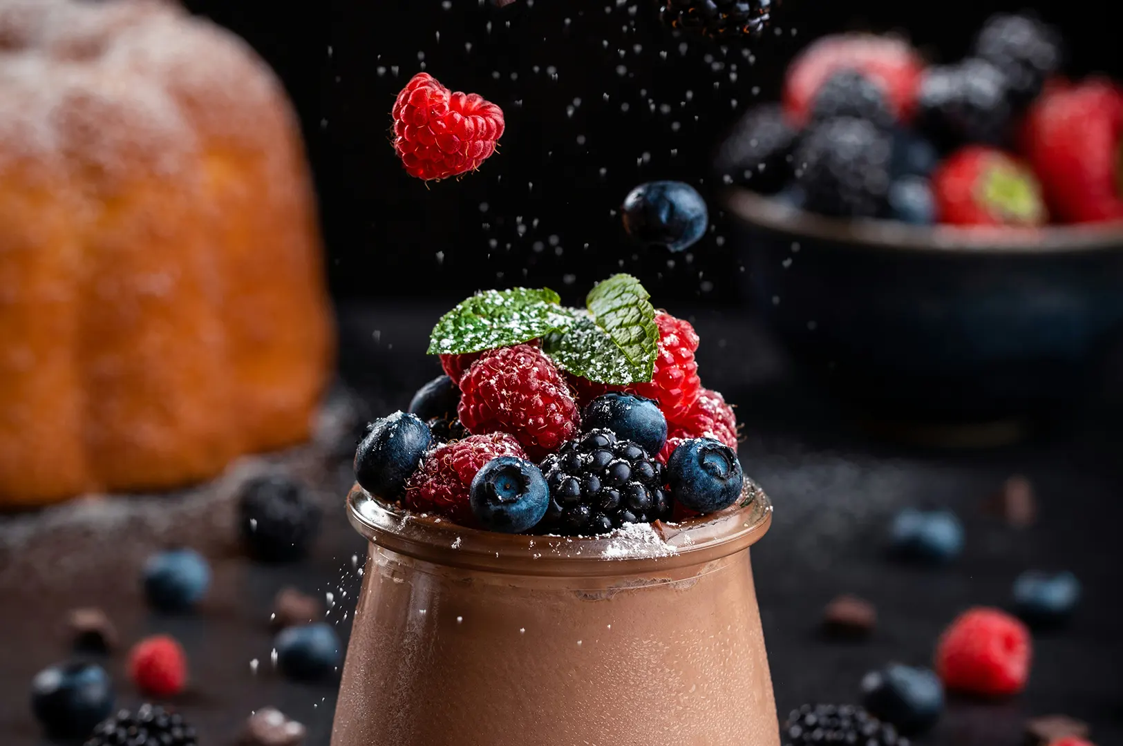 Dark moody food photography of chocolate dessert topped with fresh berries, raspberries, blueberries, and mint against a black background.