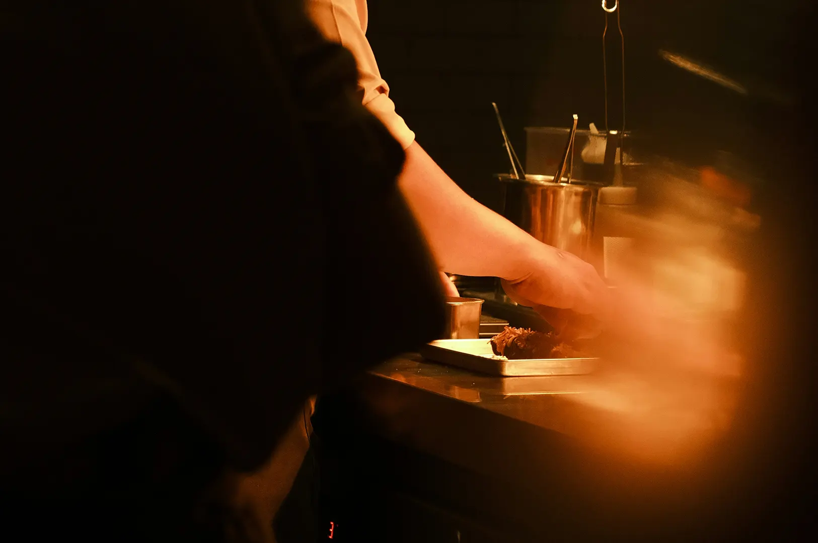 Chef plating food in a dark restaurant kitchen with staff members in the frame.