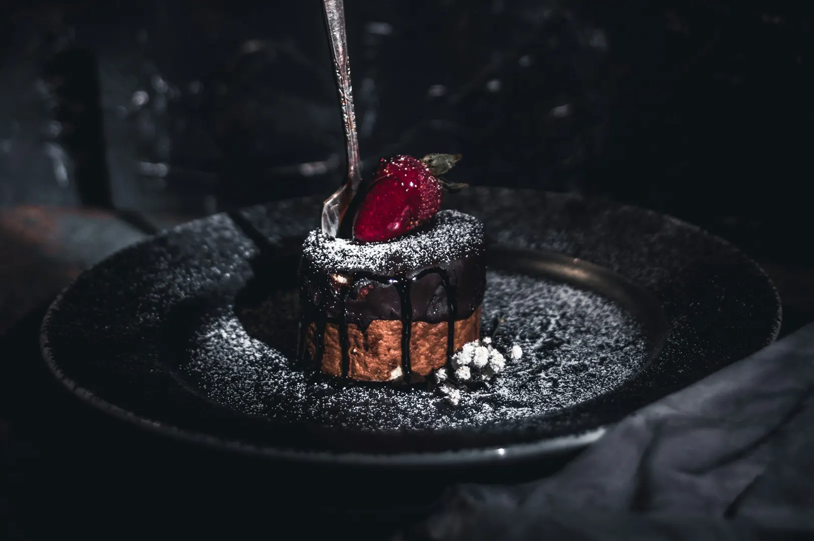 Dark moody food photography of a chocolate dessert topped with a strawberry and powdered sugar on a black plate.