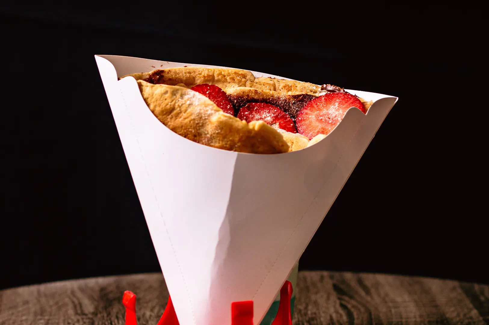 Dark mood food photography of a strawberry chocolate crepe in a paper cone container on a wooden table.