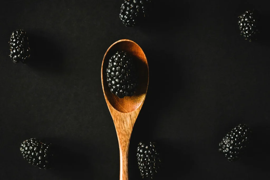 Dark moody food photography of fresh blackberries on a wooden spoon against a black background.