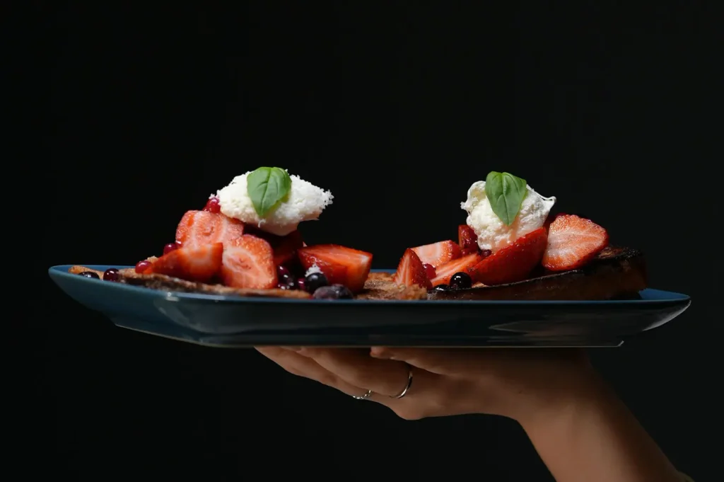 Hand holding a plate of french toast with strawberries and cream against a dark background.