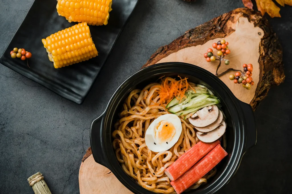 Dark aesthetic food photography of a noodle bowl and corn on a rustic tabletop.