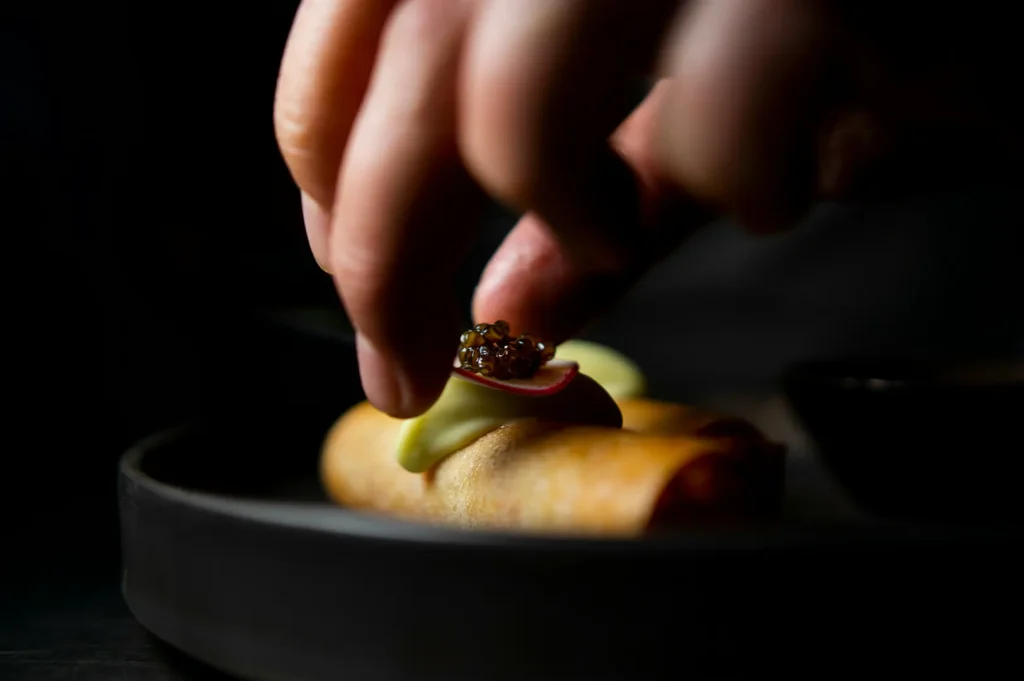 Chef plating a gourmet spring roll with caviar and cream sauce on a dark plate for restaurant food photography.