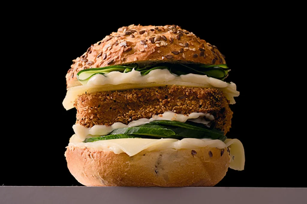 Gourmet breaded chicken burger with fresh cucumber, cheese, and mayonnaise on a dark background.