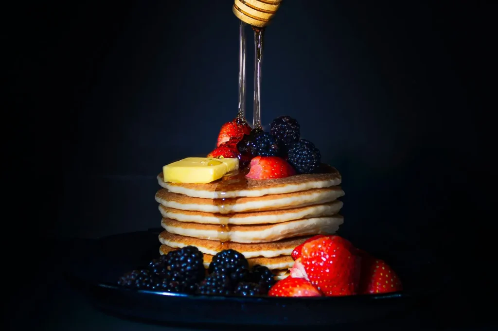 Stack of fluffy pancakes topped with butter, fresh berries, and drizzled honey on a dark background.