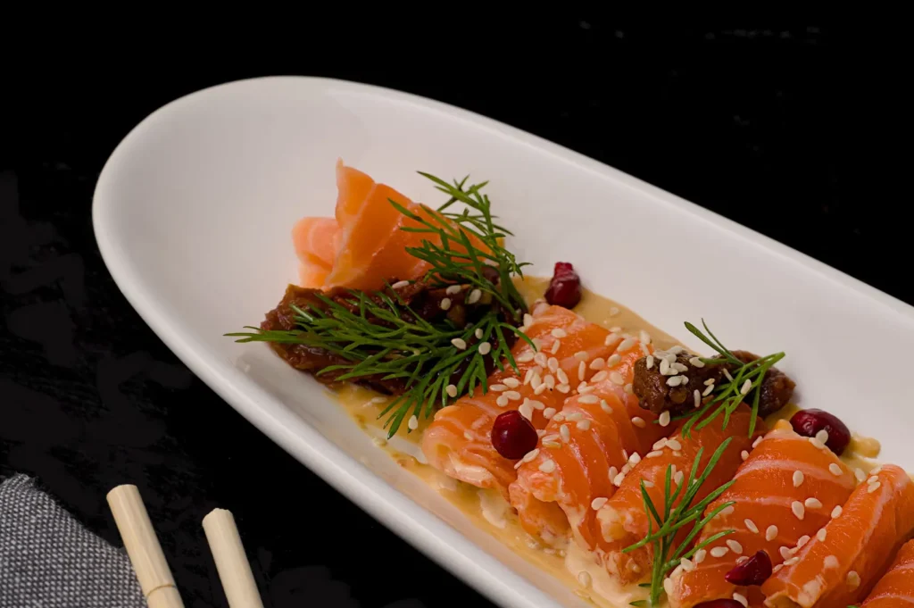 Moody food photography of sliced salmon rolls with sesame seeds on a white plate against a dark background.
