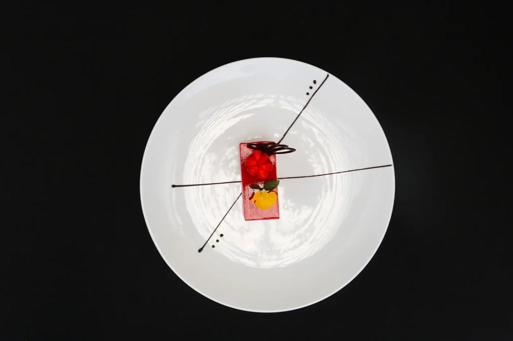 Dark moody food photography of an elegant red dessert cake artfully plated on a white plate with chocolate drizzle and black background, top view.