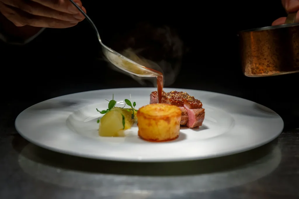 Fine dining chef pouring sauce over a gourmet dish in a restaurant kitchen with dark styling.