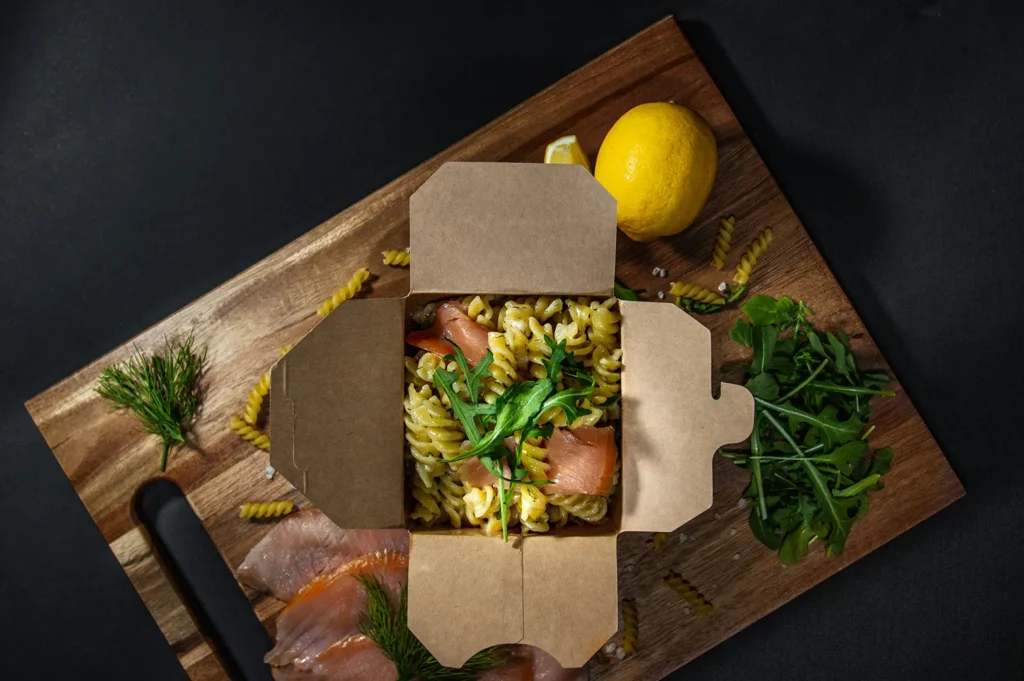 Cloud kitchen pasta takeout delivery box on a wooden board with black background.