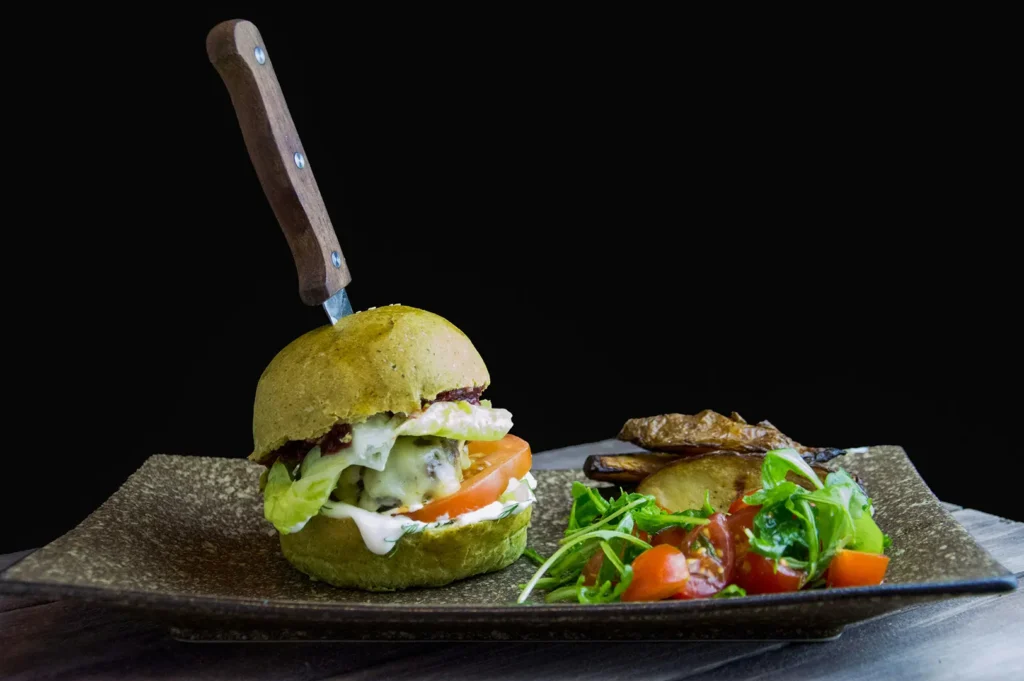 Gourmet green bun burger with a knife stuck in it, served with potato wedges and a tomato arugula salad on a black background.