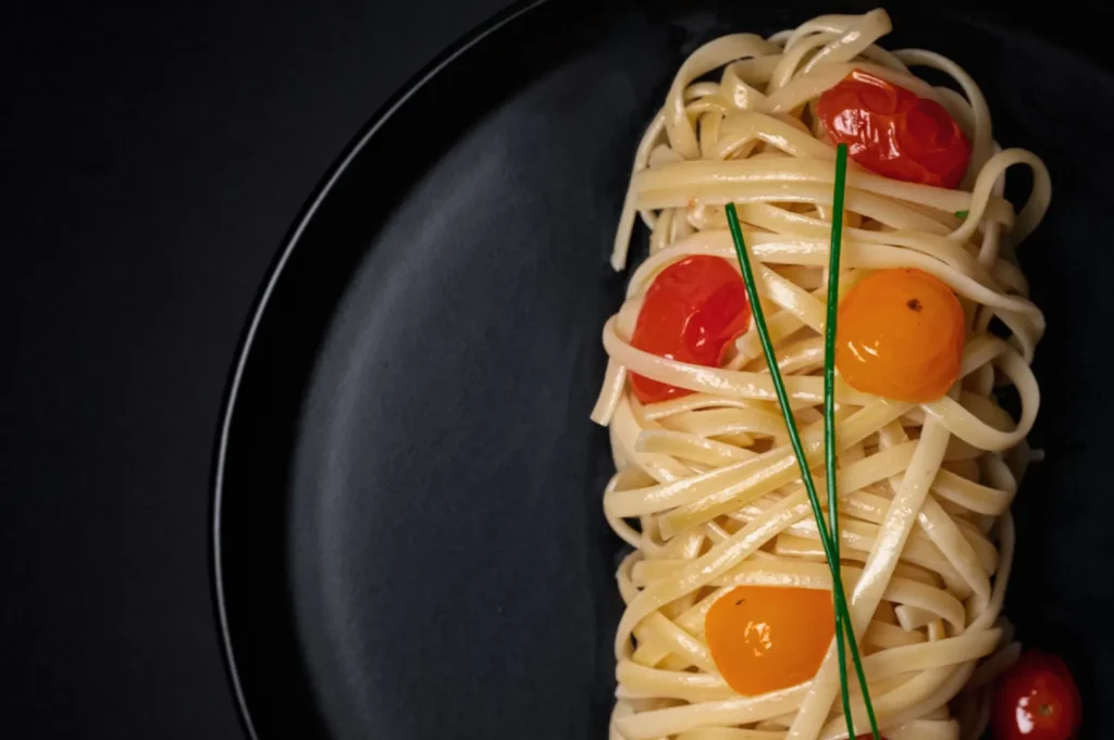 Dark moody food photography of gourmet pasta with cherry tomatoes and chives plated on a black plate.