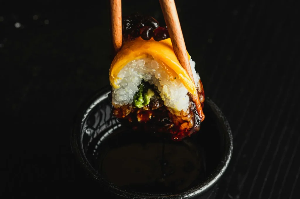 Chopsticks dipping a sushi roll with cheese and soy sauce against a dark background.