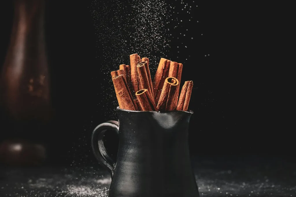 Dark moody food photography of cinnamon sticks in a black ceramic pitcher with powdered sugar dusting.