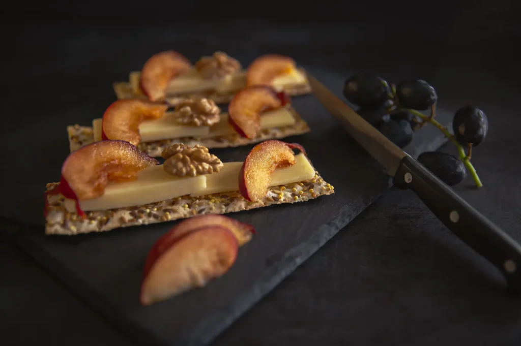 Gourmet cracker appetizers with cheese, peach slices, and walnuts served on a black slate with grapes.