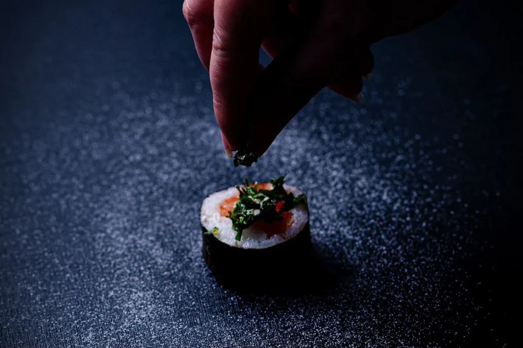 Dark moody food photography of a chef garnishing a gourmet maki sushi roll on a black textured background with copy space.