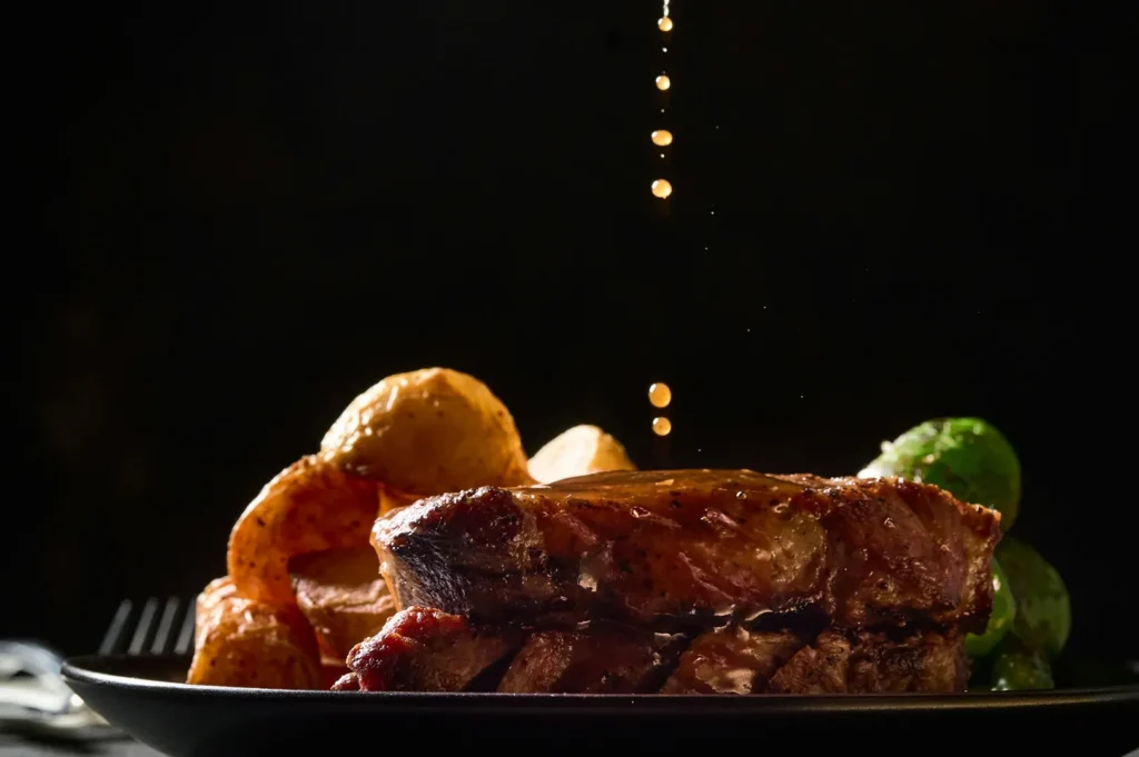 Dark-themed commercial food photography of a grilled steak meal with roasted potatoes on a black plate against a black background.