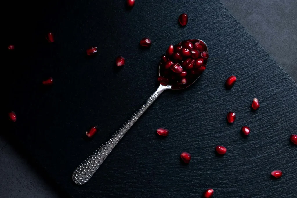 Dark food photography featuring a silver spoon filled with fresh red pomegranate arils on a black slate background.