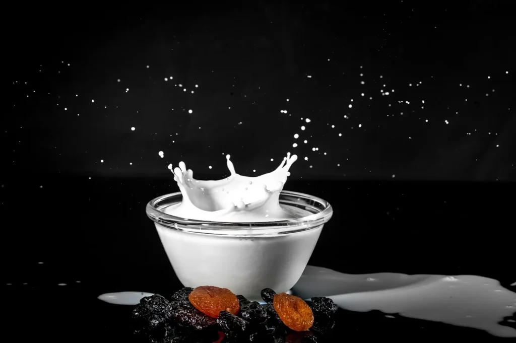Glass bowl of milk splashing with dried fruits on a dark background.