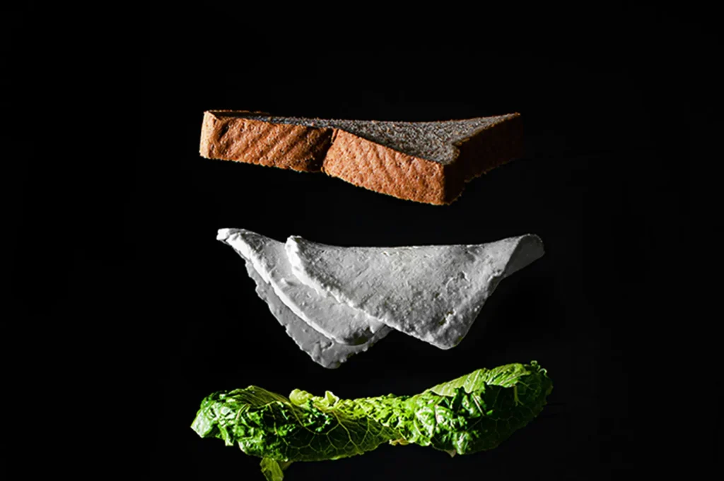 Dark moody food photography of floating sandwich ingredients including bread, cheese, and lettuce on a black background.