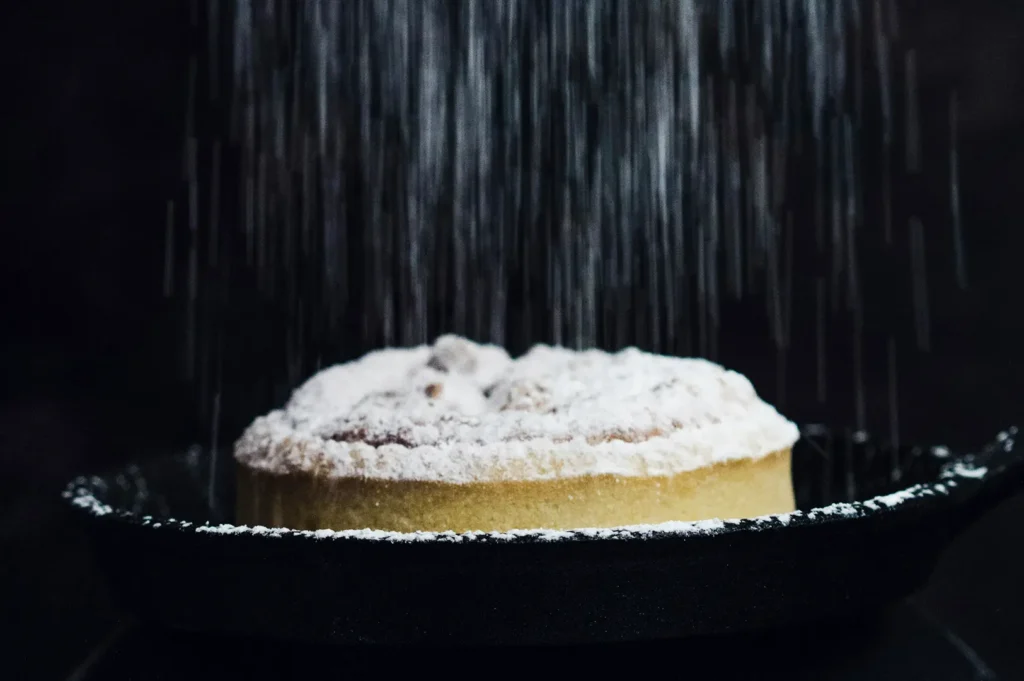 Close-up of a dark moody food photography shot showing powdered sugar falling onto a tart or pie inside a cast-iron pan.