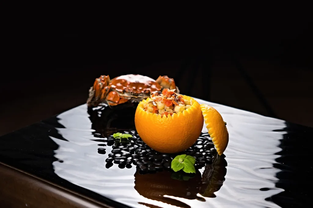 Fine dining gourmet seafood dish served in a carved orange with a whole crab on a glossy black reflective surface.