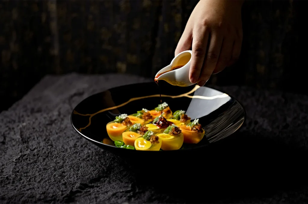 Professional dark-themed food photography of a chef pouring sauce over a plated gourmet dish.