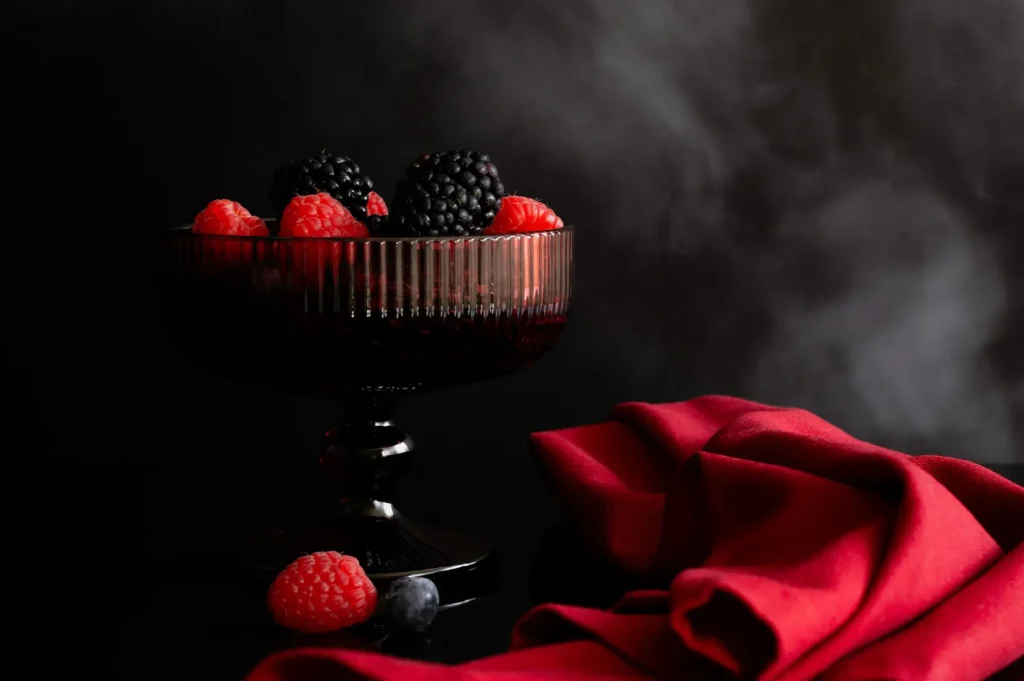 Dark moody food photography of fresh raspberries and blackberries in a glass bowl on a black table with a red cloth.