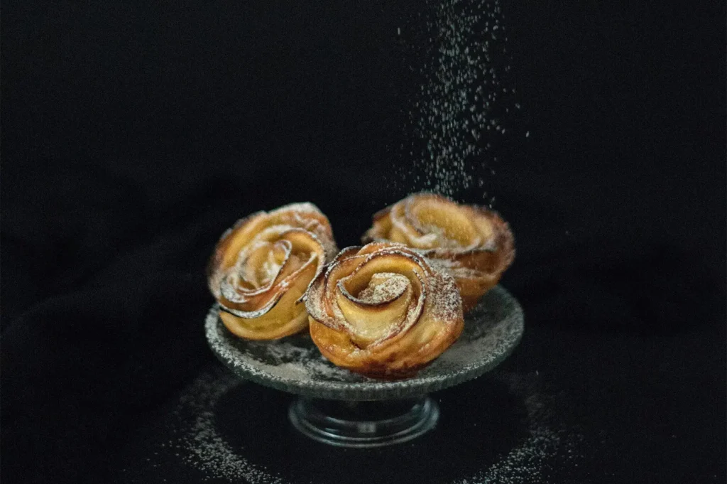 Dark moody food photography of apple pastries on a stand with powdered sugar falling, shot in RAW for a restaurant portfolio.