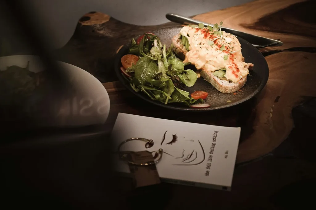 Gourmet avocado toast with scrambled eggs and side salad served on a dark plate on a rustic wooden table.
