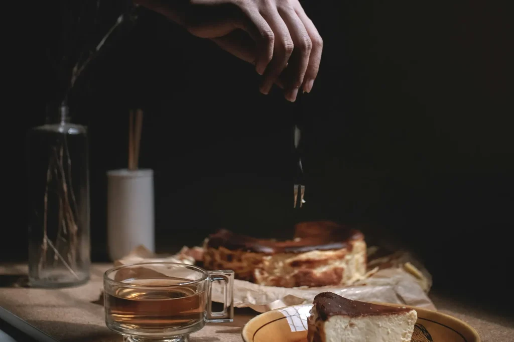 Moody dark food photography of a hand reaching for a cheesecake dessert with a dark background.
