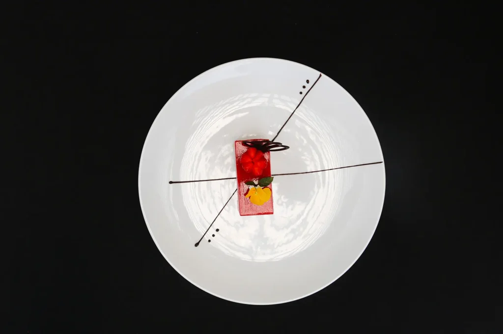 Dark moody food photography of a gourmet red dessert plated on a white plate with artistic chocolate lines against a black background.
