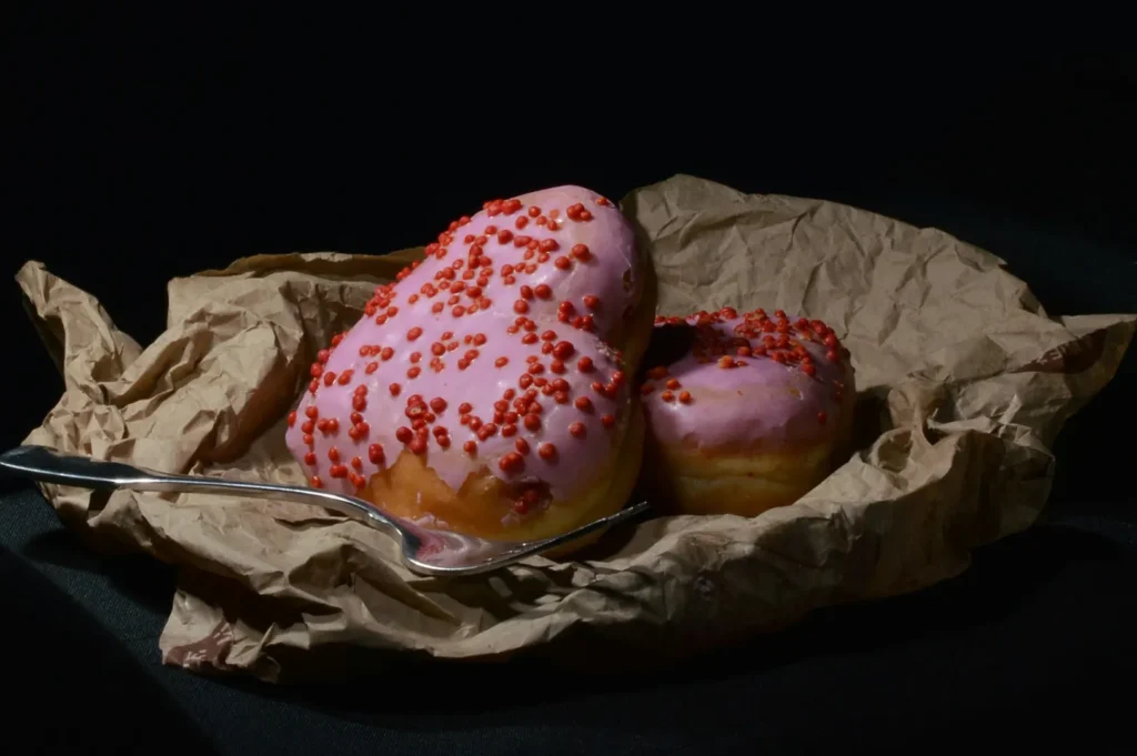 Dark moody food photography of pink glazed heart-shaped donuts with red sprinkles on brown craft paper with a silver spoon.