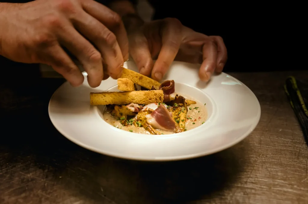 Chef plating a gourmet soup dish with breadsticks in a dark restaurant setting.