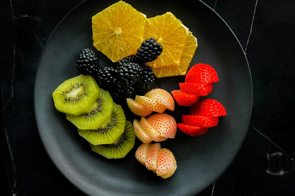 Overhead shot of sliced kiwi, oranges, blackberries, and strawberries arranged neatly on a dark plate for a restaurant menu.