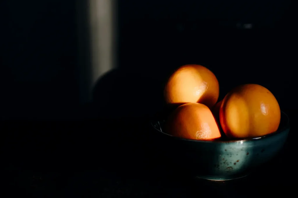 Dark moody food photography of fresh oranges in a ceramic bowl with dramatic lighting.