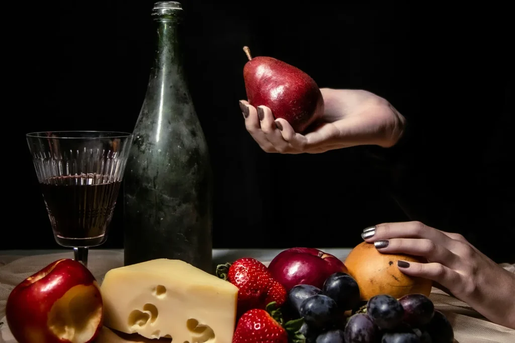 Dark moody food photography featuring a spread of fruits, cheese, wine glass, and bottle against a black background.