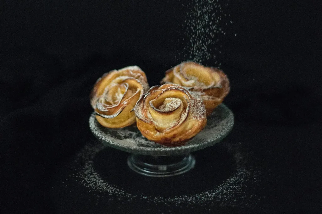 Dark moody food photography of three apple pastries shaped like roses on a small glass stand with powdered sugar being dusted over them against a black background.