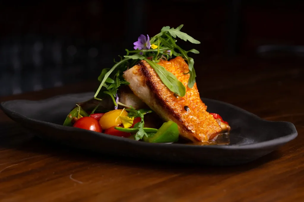 Dark moody food photography of pan-seared fish fillet with fresh vegetables and edible flowers served on a black plate.