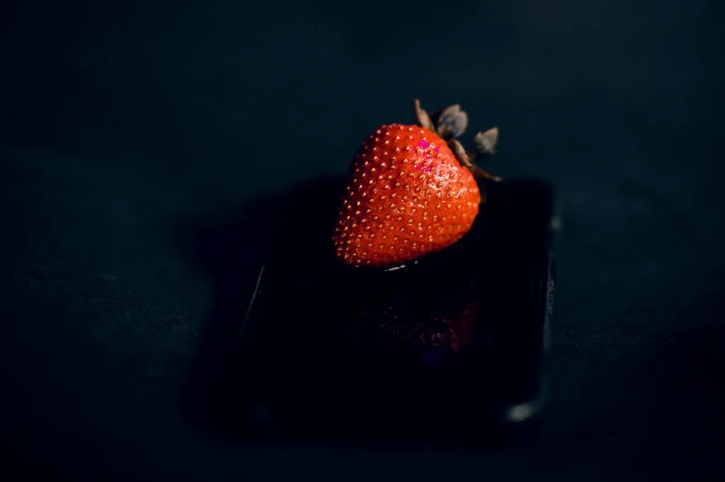 A fresh red strawberry placed on top of a black smartphone against a dark background for creative food photography.