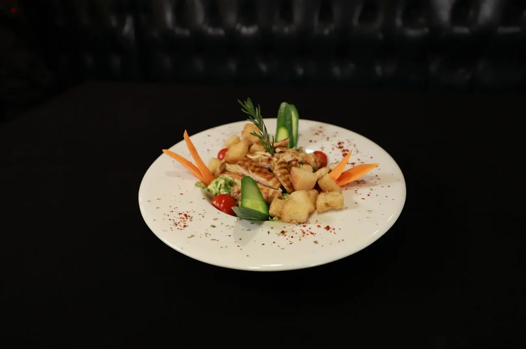 Gourmet chicken salad with croutons and fresh vegetables plated on a white dish against a black background.